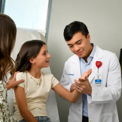 Doctor in white coat and name tag smiling and interacting with a young girl and her mother in an exam room.