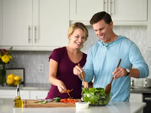 A man and woman smiling while preparing a salad together in a modern kitchen.