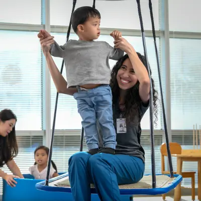 Occupational therapist assisting a young child on a therapeutic swing during a pediatric therapy session in a bright clinical room