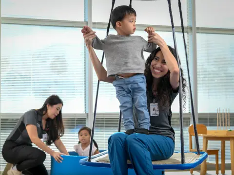 Occupational therapist assisting a young child on a therapeutic swing during a pediatric therapy session in a bright clinical room