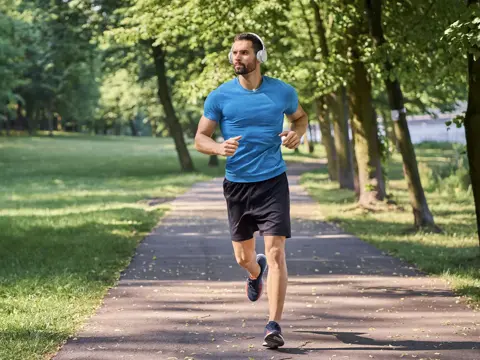 Man jogging on a tree-lined park path, wearing a blue shirt, black shorts, and over-ear headphones