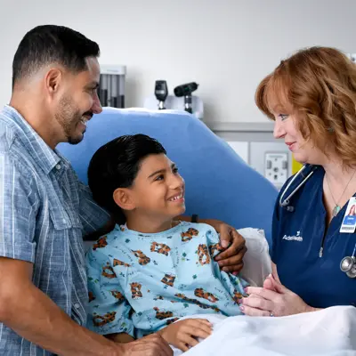 A smiling young boy in a hospital bed with his father and a nurse, all smiling.