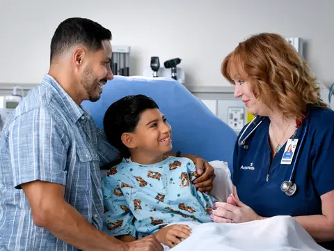 A smiling young boy in a hospital bed with his father and a nurse, all smiling.