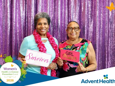 Two women holding 'Survivor' and 'Girl Power' signs at the 2026 AdventHealth Women's Health and Cancer Prevention Event against a purple sequin backdrop
