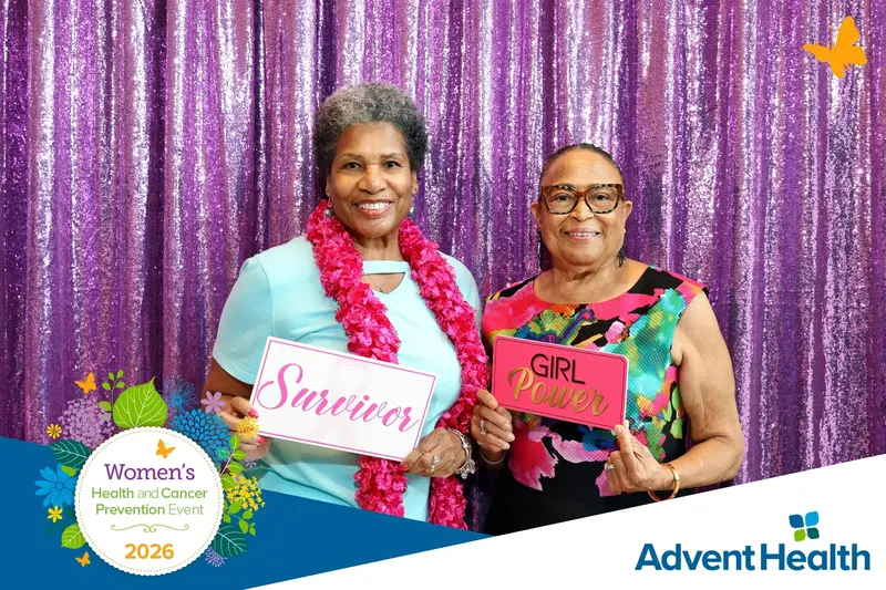 Two women holding 'Survivor' and 'Girl Power' signs at the 2026 AdventHealth Women's Health and Cancer Prevention Event against a purple sequin backdrop
