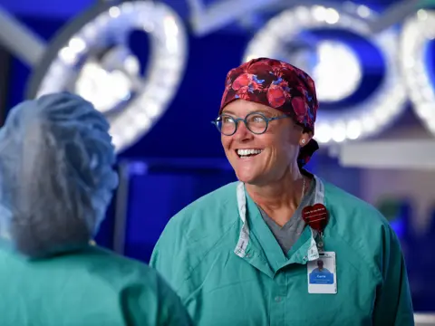 Smiling female surgeon in scrubs and surgical cap stands in operating room.