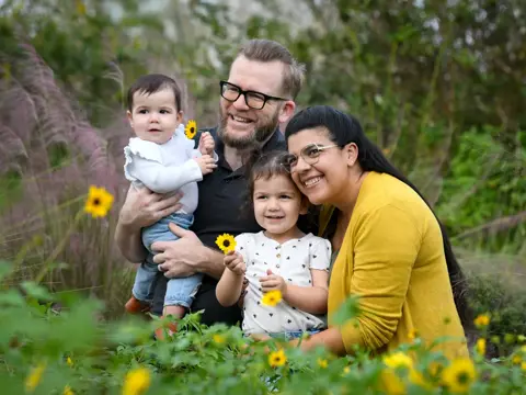 A family of four, two adults and two children, smiling and holding sunflowers in a field.