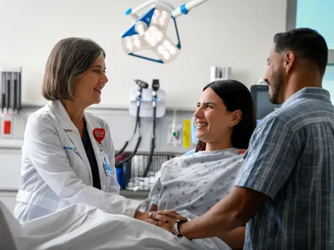 A smiling doctor and a man hold the hands of a smiling woman in a hospital bed.