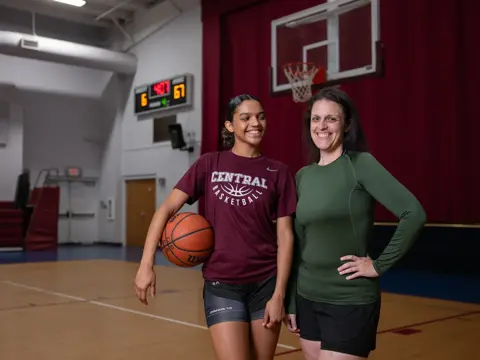 Two smiling women stand on an indoor basketball court, one in a maroon Central Basketball shirt holding a basketball, the other in a green athletic top, with a hoop and scoreboard visible in the background