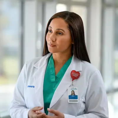 A female doctor in a white coat and green scrubs talks to a male patient in a hospital setting.