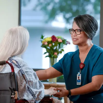 Nurse in blue scrubs holding the hand of a patient in a wheelchair, both smiling.