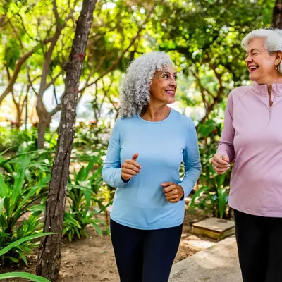 Two women with gray hair walk and smile at each other on a shaded park path, wearing long-sleeve tops and leggings.