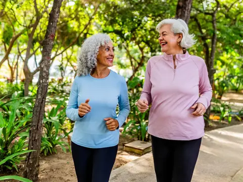 Two women with gray hair walk and smile at each other on a shaded park path, wearing long-sleeve tops and leggings.