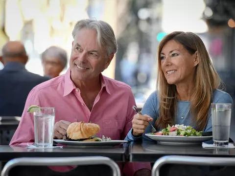 A smiling man and woman dining outdoors with plates of food and drinks.