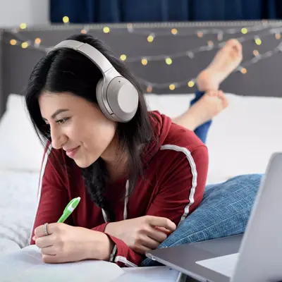 A woman wearing headphones and writing in a notebook while lying on a bed with a laptop.