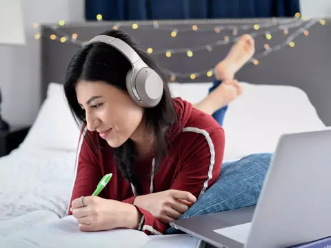 A woman wearing headphones and writing in a notebook while lying on a bed with a laptop.