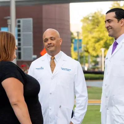 Two doctors in white lab coats and ties speak with a woman in a black shirt outside a hospital.