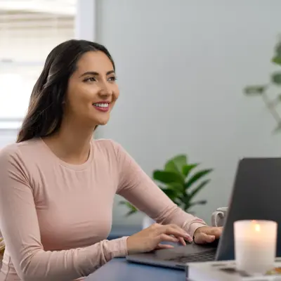 A smiling woman in a pink shirt works on her laptop at home with a candle and plants nearby.