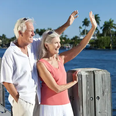 Man and woman standing together near the water, smiling and waving.
