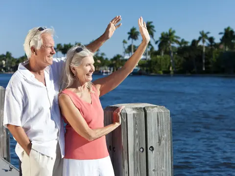Man and woman standing together near the water, smiling and waving.