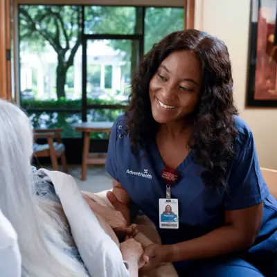 A smiling nurse in blue scrubs holds the hand of an elderly patient in a hospital bed.
