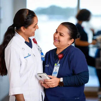 Two smiling AdventHealth employees, one in a white coat and the other in a blue uniform, stand in a hospital corridor.