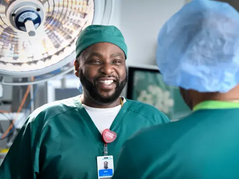 Smiling male surgeon in green scrubs and cap stands in operating room.