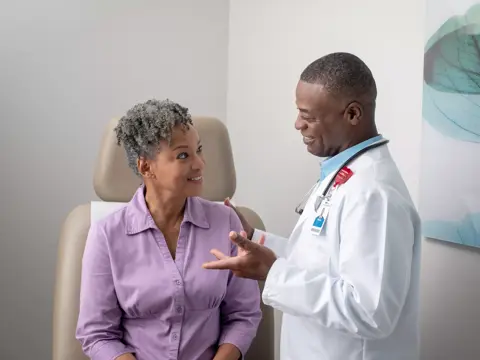 A smiling doctor in a white coat talks to a seated patient in a purple shirt.