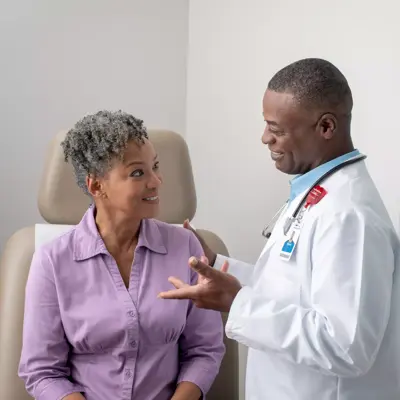 A smiling doctor in a white coat talks to a seated patient in a purple shirt.
