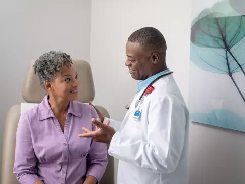A smiling doctor in a white coat talks to a seated patient in a purple shirt.
