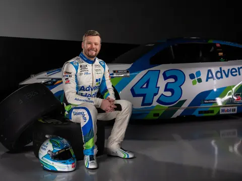 Race car driver, Erik Jones, in white and blue suit sits on stacked tires beside #43 AdventHealth NASCAR car; helmet on floor.