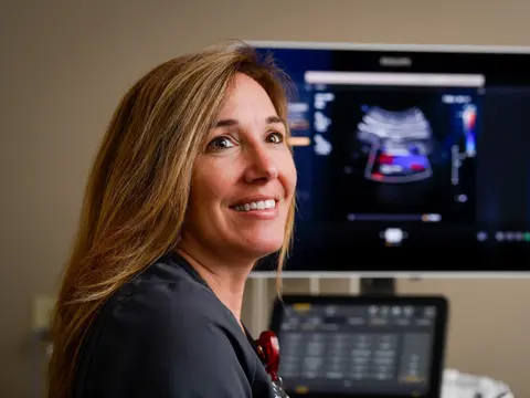 A smiling woman in a black shirt stands in front of a monitor displaying an ultrasound image.