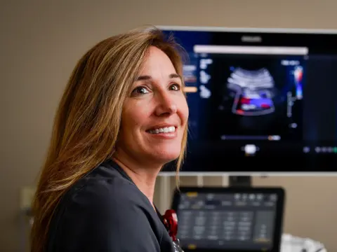 A smiling woman in a black shirt stands in front of a monitor displaying an ultrasound image.
