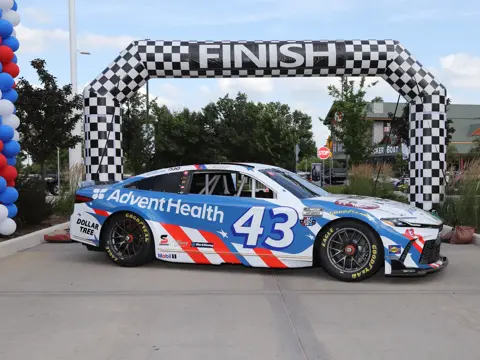 Blue and white race car with AdventHealth branding and number 43 is parked beneath a checkered finish arch at an outdoor event