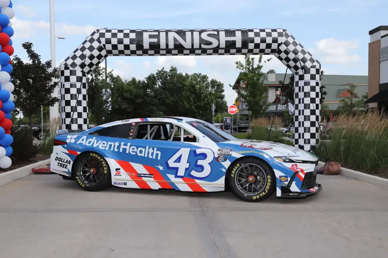 Blue and white race car with AdventHealth branding and number 43 is parked beneath a checkered finish arch at an outdoor event