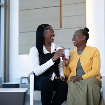 Two women sitting and laughing together while holding coffee cups in a waiting room.