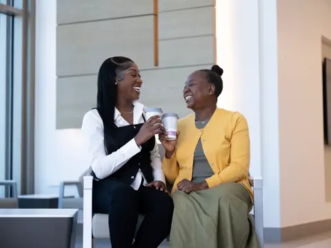 Two women sitting and laughing together while holding coffee cups in a waiting room.