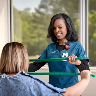 A smiling physical therapist assisting a patient with a resistance band exercise in a bright room.
