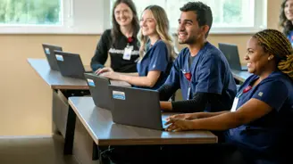 Healthcare professionals in navy scrubs sit at desks using laptops during a nurse residency training session in a bright classroom, smiling and engaged in learning.