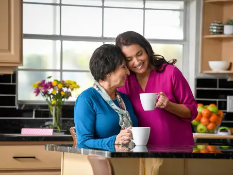 Two women in a kitchen, one holding a coffee cup, smiling and engaged in conversation.