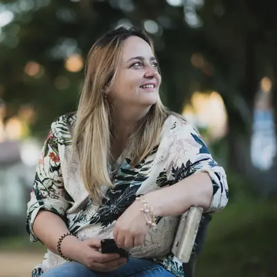 Smiling woman with long blonde hair sitting outdoors in a park, wearing a floral blouse, holding a smartphone and looking upward with a hopeful expression