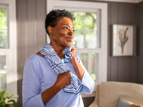 A woman in a blue shirt and striped scarf stands in a living room, looking thoughtful.