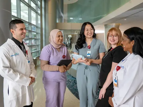 Five diverse medical professionals in scrubs and white coats standing together in a modern hospital lobby, smiling and engaged in conversation