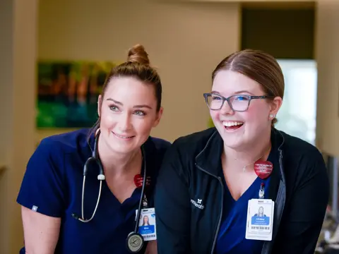 Two smiling nurses in blue uniforms, one with a stethoscope, stand together in a medical office.