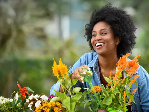 A smiling woman in a denim jacket watering vibrant flowers in a garden.