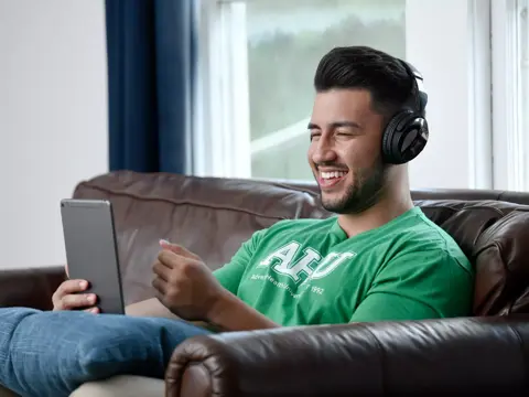 A smiling man wearing headphones and a green shirt with AHU logo, sitting on a couch and holding a tablet.