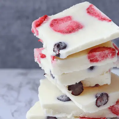Stack of white frozen yogurt bark squares with sliced strawberries and blueberries on a light marble surface.