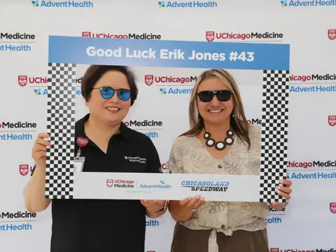 Two women smile while holding a racing-themed photo frame that reads 'Good Luck Erik Jones #43' in front of a UChicago Medicine and AdventHealth event backdrop with checkered flag patterns