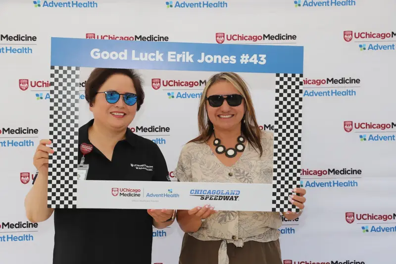 Two women smile while holding a racing-themed photo frame that reads 'Good Luck Erik Jones #43' in front of a UChicago Medicine and AdventHealth event backdrop with checkered flag patterns