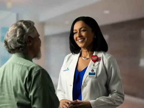 A smiling female doctor in a white coat talks to a male patient in a green shirt.